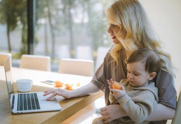 eine Frau guckt auf den Bildschirm des Laptops, auf ihrem Schoß sitzt ein Kleinkind, das eine Mandarine in der Hand hält