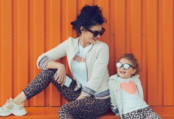 Mutter und Tochter vor oranger Blechwand, beide tragen Hosen mit Leoprint und Sonnenbrille