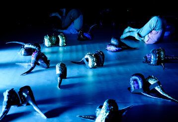 My tail and I Tanzperformance, blaue Beleuchtung, zwei Kinder liegen mit den beinem über dem Kopf auf der Bühne, glitzernde Dino-Schwänze liegen herum