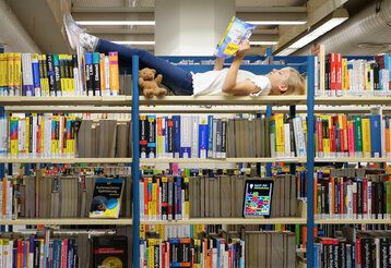 Ein Mädchen liegt auf einem Bücherregal einer Bibliothek und liest, ein Teddybär sitzt auch im Regal