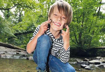 Ein Junge sitzt an einem bach und schmiert sich Matsch ins Gesicht