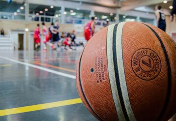 Im Vordergrund ein großer Baskeltball, im Hintergrund eine Sporthalle mit Sportlern