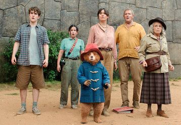 Szene aus dem Film Paddington in Peru, Paddington Bär und fünf Leute (Junge, Mädchen, Frau, Mann, Oma) stehen vor einer Mauer