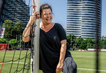 Petra Berg steht auf einem Fußballplatz und lehnt sich an ein Tor. Im Hintergrund Hochhäuser.