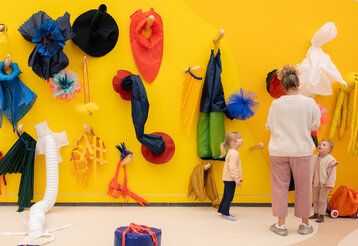 Raum mit gelber Wand und bunten Stoff-Objekten, eine Frau und zwei Kinder stehen zusammen