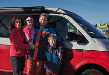 Familie (Vater, Mutter, Kind und Baby) steht vor VW-Bus, im Hintergrung Zirkuszelt, Wintersonne mit harten Schlagschatten