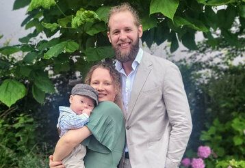 Porträt der Familie Passen, Mutter trägt Baby auf dem Arm, festlich gekleidet, im Hintergund  Pflanzen