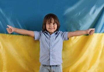 Studio Porträt von einem kleinen Jungen der vor einer ukrainischen Flagge steht und lächelnd seine Daumen hebt