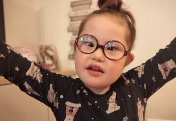 kleines Mädchen mit Down Syndrom und Brille guckt in die Kamera