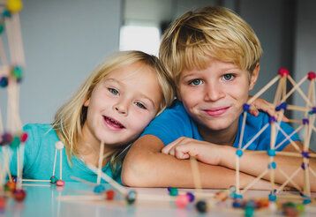 Porträt von zwei Kindern beim Basteln, im Vordergrund unscharf: Gebilde aus Holzstäbchen und Knete