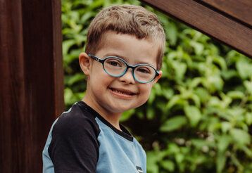 Porträt eines Jungen mit Down-Syndrom, er trägt eine Brille und lächelt in die Kamera, im Hintergrund Gebüsch
