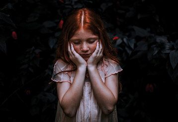 ein Mädchen steht vor einem sehr dunklem Hintergrund, es hat die Augen geschlossen und hält sich die Hände an die Wangen, im hintergrund erkennt man Blätter einer Pflanze