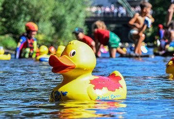 Ein Quietscheentchen schwimmt in der Wupper, im Hintergrund weitere Entchen und Kinder, die im Wasser spielen