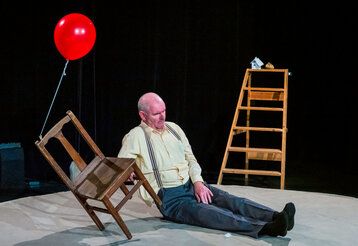 Ein Mann mit Glatze und Hosenträgern sitzt auf einer Bühne zwischen einem Stuhl und einer Holzleiter.