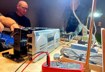 aufgeschraubter Ghettoblaster, Kabel  und Gerätschaften auf einem Tisch, drei Männer beim Reparieren