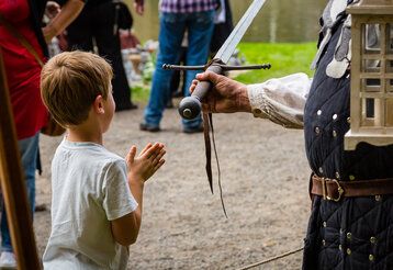 ein kleiner Junge steht bei einem Ritter, der ihm ein Schwert reicht