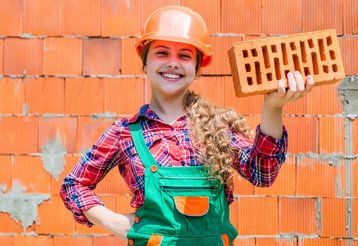 Mädchen mit Bauhelm vor einer gemauerten Wand auf einer Baustelle.