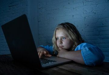 Mädchen sitz in einem dunlen Zimmer und guckt auf den leuchtenden Bildschirm eines Laptops