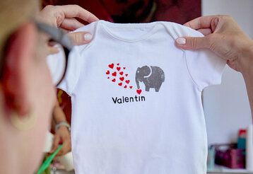Frau hält selbstgestalteten Babybody in den Händen, auf dem Body ist ein Elefant mit roten Herzchen abgebildet und Schrift: Valentin