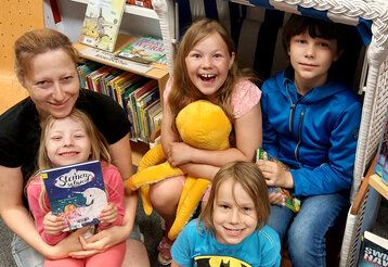 Gruppenbild: eine Frau und vier Kinder sitzen in einer Stadtbücherei auf dem Boden, ein Kind hat ein Buch in der Hand, ein anderes ein Stofftier