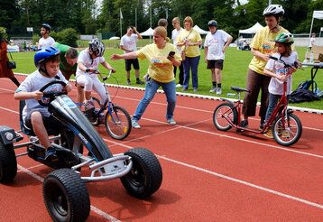 Kinder mit sonderpädagogischem Unterstützungsbedarf fahren mit verschiedenen Fahrzeugen auf einer Tartanbahn, Erwachsene gucken zu und helfen