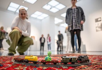 Spielzeugeisenbahn auf einem Teppich bei der Hans-Peter Feldmann-Ausstellung im Kunstpalast Düsseldorf, Besucherinnen, unscharf im Hintergrund, betrachten die Bahn