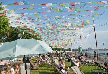 Stadtstrand Robert Lehr Ufer, Leute liegen auf Liegestühlen auf der Wiese, tibetische Gebetsfahnen flattern im Wind, Rhein mit Blick auf Kniebrücke im Hintergrund
