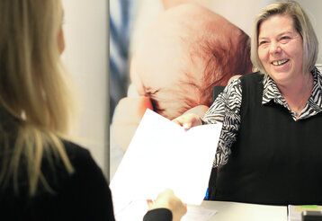 Standesbeamte überreicht lachend ein Dokument, sie sitzt am Schreibtisch, eine Frau sitzt ihr gegenüber und nimmt das Dokument an