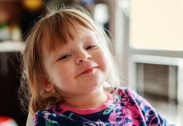 Kleines Mädchen guckt frech grinsend in die Kamera, im HIntergrund unscharfer Raum