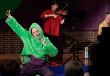 eine Frau mit einem grünen Teddy-Pullover kniet auf der Bühne und streckt den Arm hoch, im Hintergrund: eine Geigenspielerin und ein Mann am Klavier
