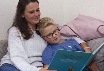 Mutter und Sohn sitzen auf dem Sofa. Das Kind liegt im Arm der Mutter. Die Mutter liest aus einem Buch vor. Perspektive von vorne.