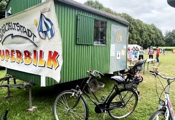Bauwagen mit einem Banner, auf dem steht: Kinderstadt Superbilk, auf einer Wiese, im Hintergrund Leute, im Vordergrund Fahrräder