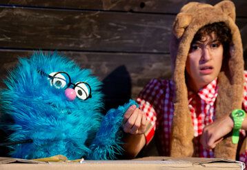 Szene aus Das Klugscheißerchen, ein blaues Plüschmonster sitzt zusammen mit einer Person im Karohemd in einer Hütte, die beiden halten Händchen