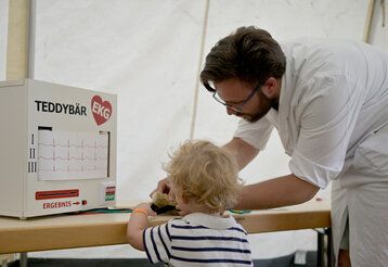 Arzt untersucht einen Teddybären, kleines Kind schaut zu