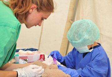 Kind in Arztkittel behandelt kranken Teddy, eine Frau assistiert