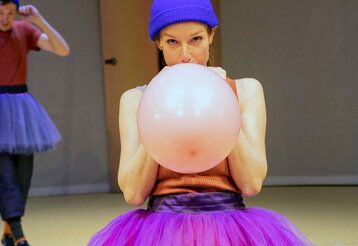 Schauspielerin im Tutu bläst Luftballon auf, Schauspieler im Tutu im Hintergrund hält sich die Ohren zu