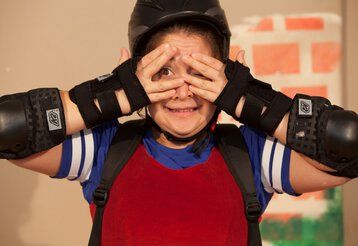 Ein Mädchen in Rollschuh- Schutzmontur hält sich die Hände vor ihr Gesicht und blinzelt ängstlich zwischen ihren Fingern hindurch.