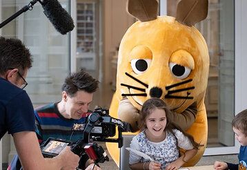 Die Maus steht hinter einem lachenden Mädchen, ein Junge steht daneben und ein Kamerateam ist dabei