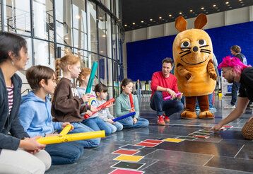 Kinder sitzen mit André Gatzke auf dem Fußboden und unterhalten sich. Die Mais steht daneben. Ein Mann legt bunte Karten auf den Boden.