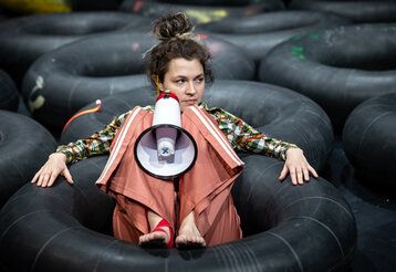 Frau sitzt mit einem Megaphone in einem schwarzen Reifen, um sie herum weitere schwarze Reifen