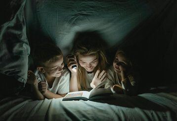 Mutter liest mit zwei Kindern unter der Bettdecke mit der Taschenlampe ein Buch