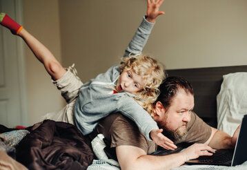 Vater liegt auf Bett mit Laptop, Kind stolpert über Vater