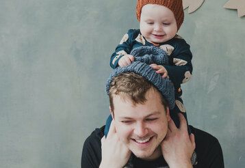 Junger Vater trägt Baby auf Schultern, Baby zieht Vater die Mütze aus