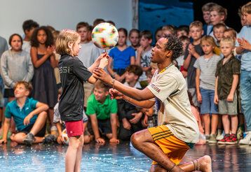 ein Mädchen balanciert einen Fußball mit der Hand, ein Mann hilft ihr dabei, im Hintergrund gucken andere Kinder zu