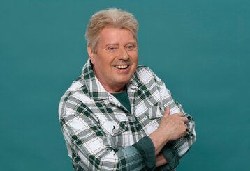 Volker Rosin steht mit verschränkten Armen vor blaugrünem Hintergrund und lacht in die Kamera