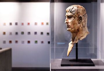 alte römische Büste in einer Glasvitrine, im Hintergrund kleine Bilder an der Wand, unscharf