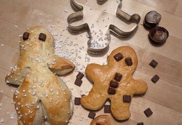 zwei selbstgebackene Weckmänner liegen auf einer Arbeitsplatte, daneben liegt ein Ausstechförmchen, Haselnüsse, Schokoflocken und grober Zucker