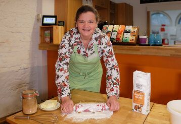 Frau mit Schürze rollt Teig mit Nudelholz aus, Backutensilien auf dem Tisch, im Hintergrund eine Küche mit Theke