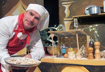 Mann mit Kochmütze und Schürze zeigt einen Teller mit Keksen, hinter ihm eine Küchen-Kulisse
