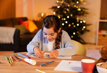 ein Mädchen klebt eine Briefmarke auf ein Papier, im Hintergrund Wohnzimmer mit Weihnachtsbaum